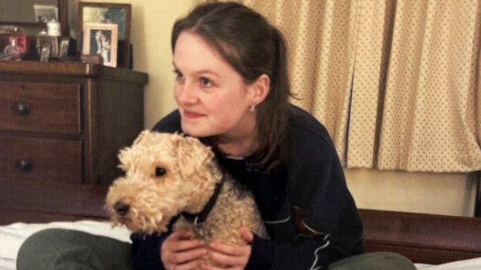 A woman in a black top and grey trousers, sitting on a bed and hugging a blonde lakeland terrier dog