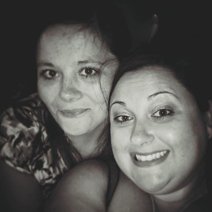 A black-and-white selfie-style photo showing the heads and shoulders of two smiling women whose faces are touching