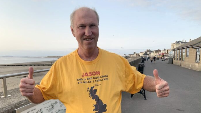 Jason Vickers in a yellow marathon t shirt as he trains in Burnham. He has both thumbs up as he smiles at the camera. In the background, you can see the beach road in Burnham.