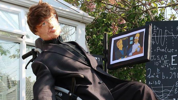 A scarecrow of Stephen Hawking. It has brunette hair, glasses and a black suit. It is in a wheelchair and sat next to a chalkboard full of equations