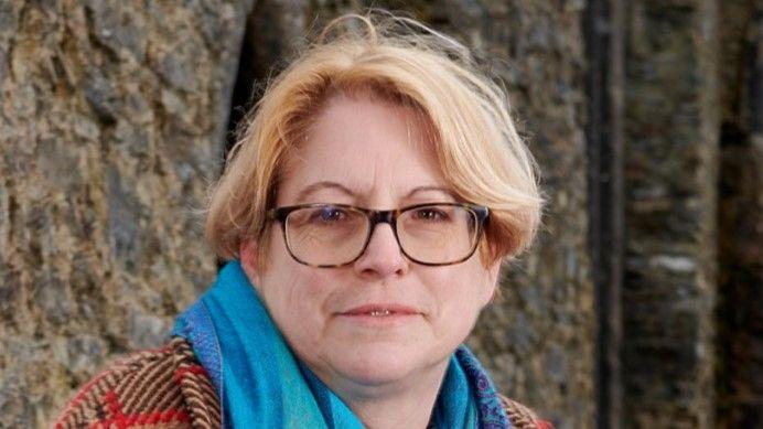 Prof Marion Gibson, sitting outside a historic stone building. She has fair, centre-parted hair and is wearing tortoiseshell glasses and a turquoise scarf over a thick red, black and brown checked coat.