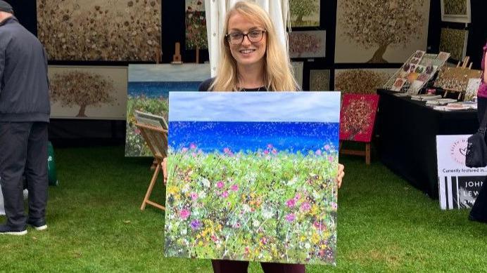 Artist Sara Otter holding up her artwork in front of a marquee filled with other paintings