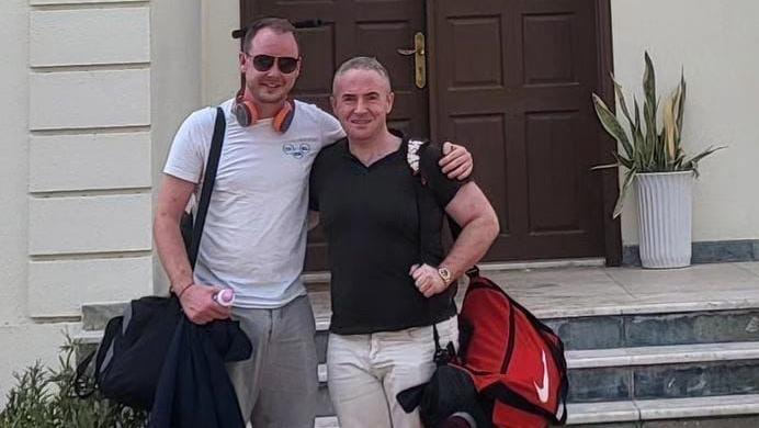 Craig Ballentine with his friend Sean Morgan standing with their arms around each other.  Craig is wearing sunglasses, headphones, a white T-shirt and grey trousers.  Sean is wearing a black T-shirt and light coloured trousers.  They are both smiling and carrying shoulder bags. 