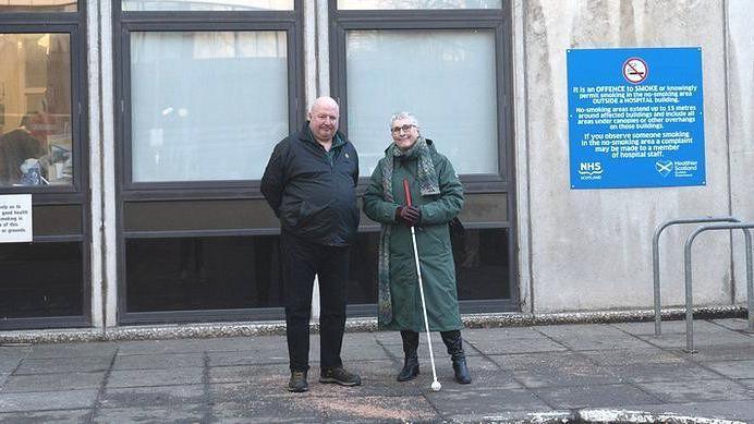 Iain Young is standing outside the hospital with a friend. He is in a black jacket and black trousers and he is resting his hands behind his back. His female friend is in a long green winter coat and has a white cane.