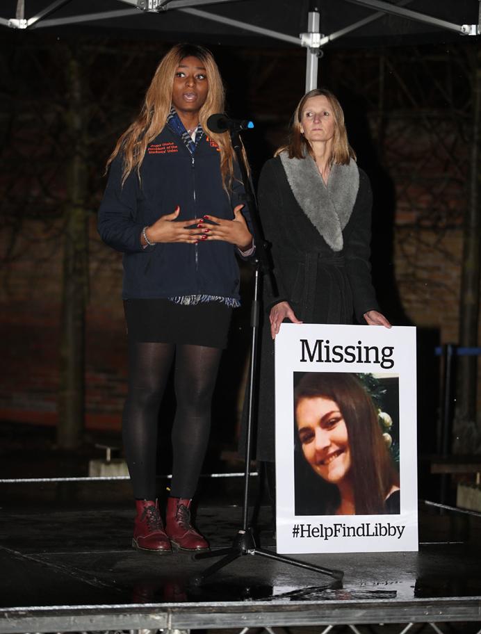 Two women on a stage with one holding a missing Libby Squire placard