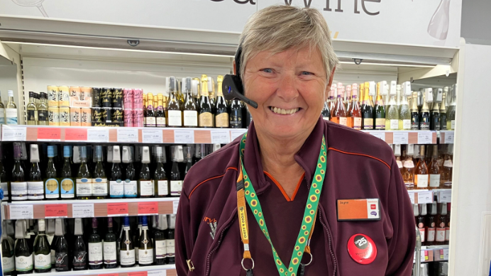 Jane Green wearing her burgundy Sainsbury uniform and earpiece with bottles of wine for sale in the background.