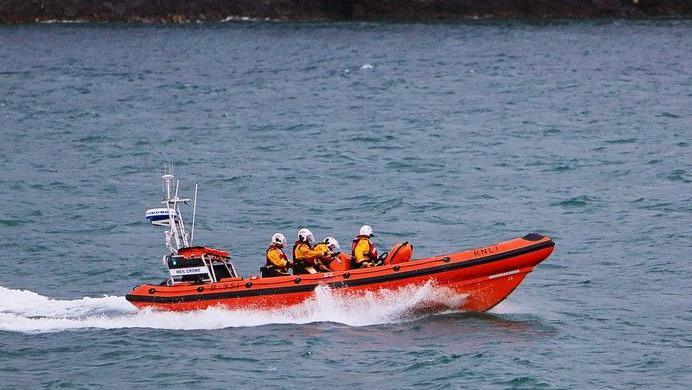 The new RNLI Lifeboat being delivered, it is a bright orange vessel, with people sat in bright yellow jackets and white helmets.
