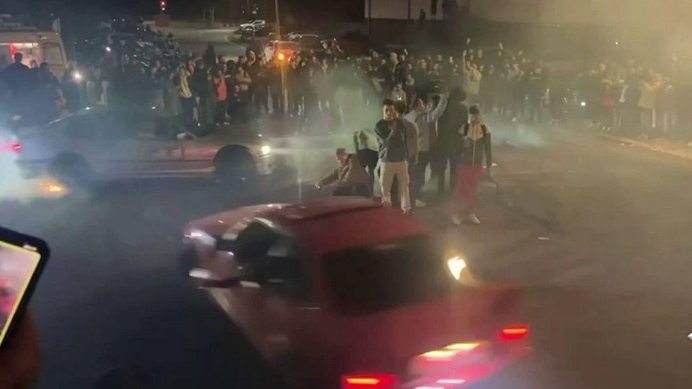 A police photograph of a night-time scene with dozens of people gathered together cheering on drivers in two cars.