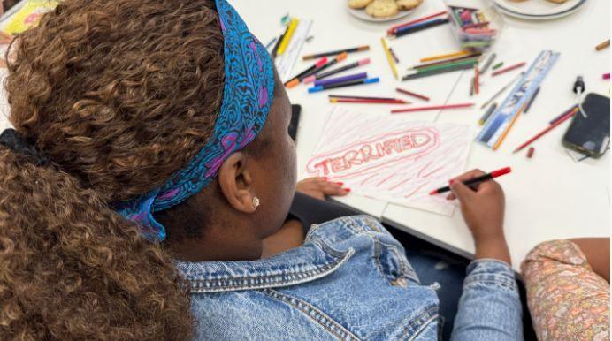 Amina, who has thick dark hair swept back with a blue bandana, illustrates a piece of paper on which she has written the word "terrified" in large red letters