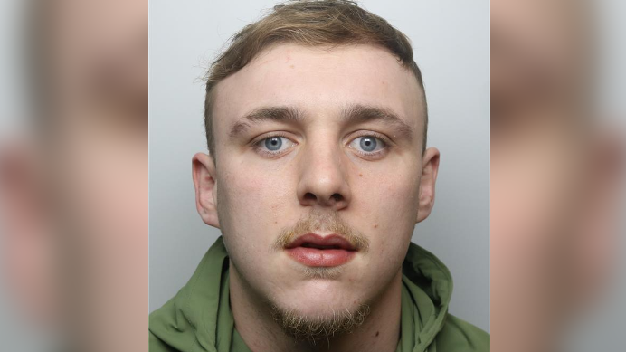Mugshot of Braden Lynam, who has blue eyes and blond hair. He is wearing a green top and has a slight facial hair.