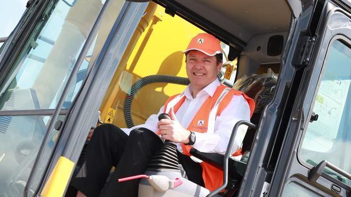 Jeremy Fish wearing an orange hi-vis vest and cap while sitting in a yellow digger