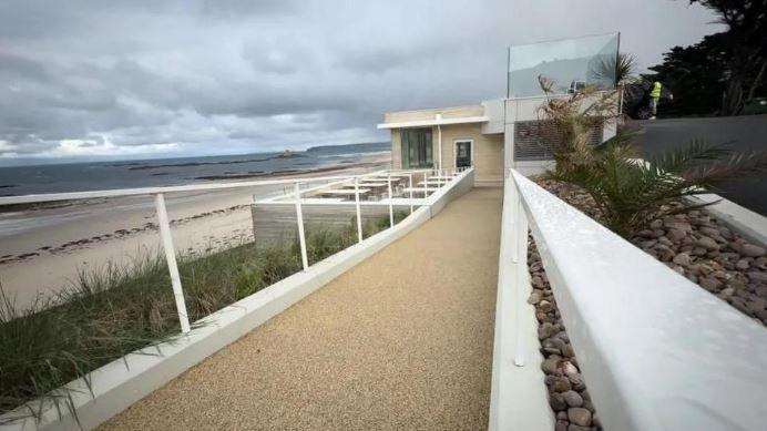 A pathway leads down to a beachside building with tables  outside and to the left a stretch of white sand bordered by the sea.