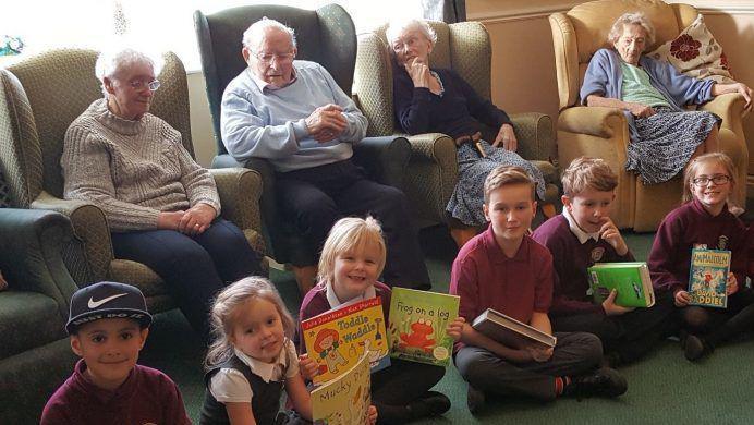 In the foreground there are six smiling primary age children, sat crossed leg holding colourful picture books. Behind them are 4 older residents from Deangate Care Home in Mapplewell, Barnsley.