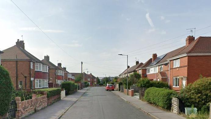A suburban street lined either side with red brick local authority-type properties. Each has a small front garden abutting the empty road, most feature priver hedges.