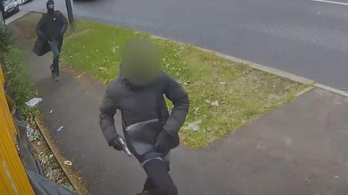 Teenager (with face blurred) dressed in black jacket and jogging bottoms holds a silver pistol as he runs down the street