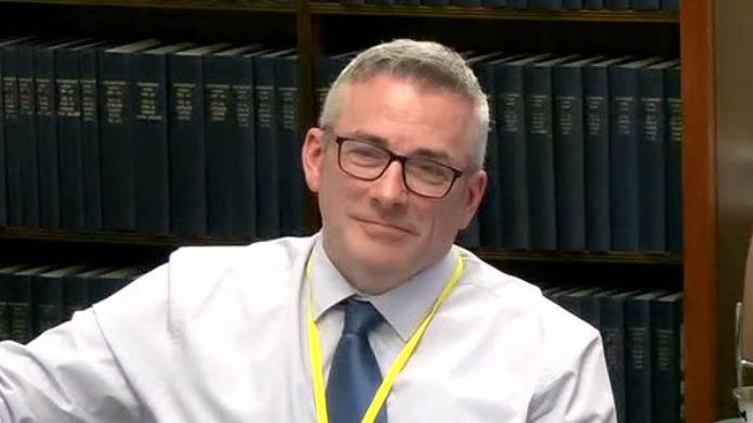 Mr Henderson with short grey hair and black square glasses. He is wearing a white shirt and blue tie.