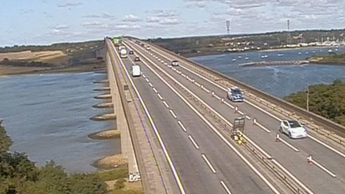 Four-lane bridge with narrow central reservation. There are cars and vans with gaps between them, There are pontoon supports visible on the waterline. There hedges on both sides of the bridge in the foreground, while fields and buildings can be seen in the background.