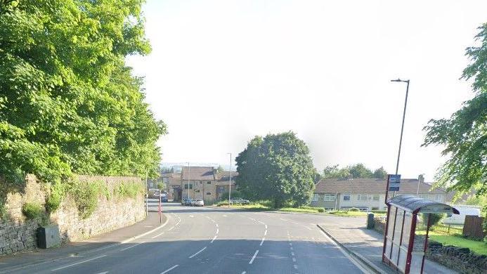 A wide suburban road on a sunny day. Leafy trees can be seen, along with a bus stop, a street light, and in the far distance, some houses and cars.