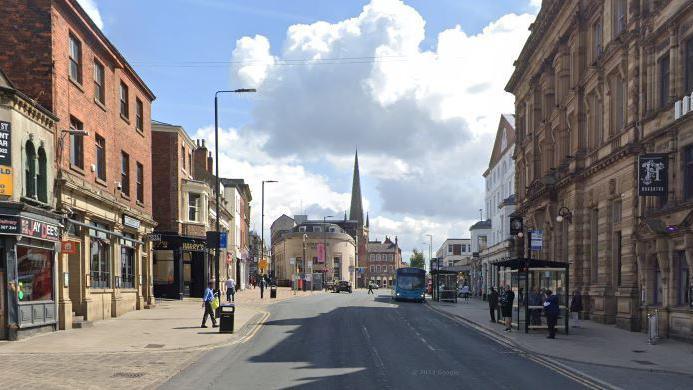 A high street in Wakefield, West Yorkshire, with stone buildings on either side and people walking on the pavements. There is a bus stop on one side with people waiting for a bus.
