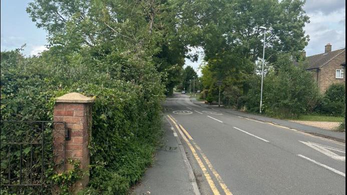 A picture of a road with double yellow lines on either side. On the left is a brick pillar with a metal gate attached. A path is next to the road and has overgrown bushes and foliage covering part of it. 