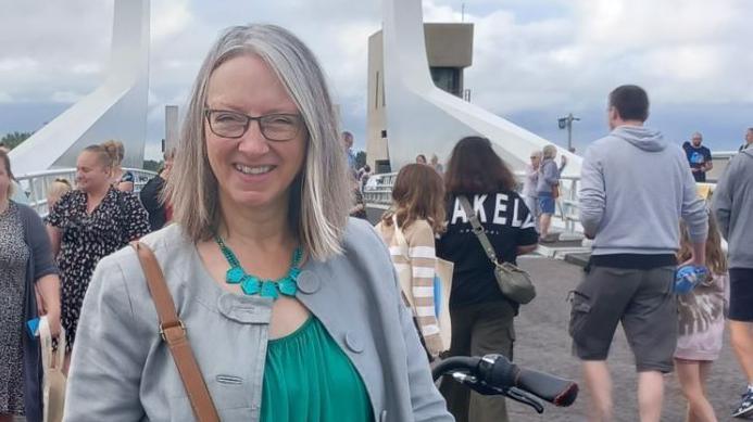 Caroline Topping is smiling at the camera and standing near the Gull Wing bridge, a white-coloured structure in Lowestoft.  She is wearing a grey jacket, turquoise top and turquoise necklace. There are people walking behind her.