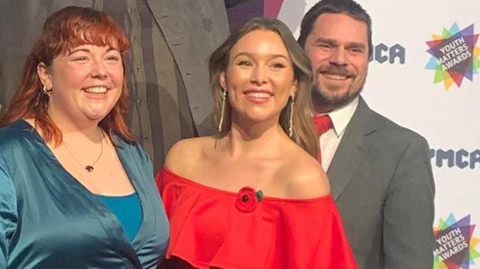 A group of people smartly dressed stand on a stage. Two women wearing dresses stand next to each other and smile. Three men dressed in suits are stood behind them. A YMCA Youth Matters Awards backdrop is behind the group.