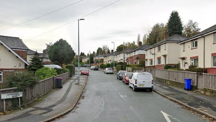 Houses are either side of a road. A sign on the left hand side says Summerville Road. There are seven cars parked along the road, along with a van.