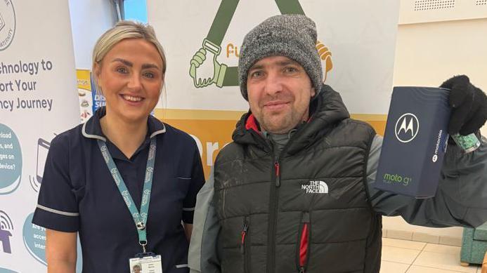 Head and shoulder shot of a man and a woman. The woman - whose blonde hair is tied back and is smiling, is wearing a dark blue hospital uniform and a lanyard around her neck. The man, to her left is wearing a puffed black gilet, and a grey beanie hat. He is smiling and holding aloft a box containing a mobile phone.