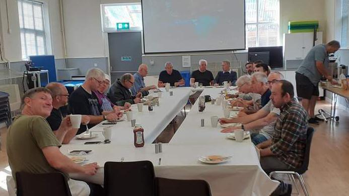 A group of men sit around a large table inside a hall eating breakfast. A man closest to the camera is smiling while the others area engaged in conversation and eating.