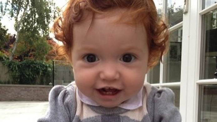 A toddler with ginger curly hair grins at the camera while sat in a garden. She is wearing a grey jumper with red hearts on