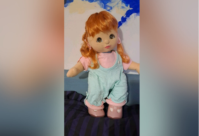 The vintage red-haired My Girl Doll that was donated to Sobell House hospice and was sold for £310 to a buyer in Italy. It is propped up on what looks like a soft service against a wall. The doll is dressed in light blue jumpsuit, a pink T-shirt and pink shoes.