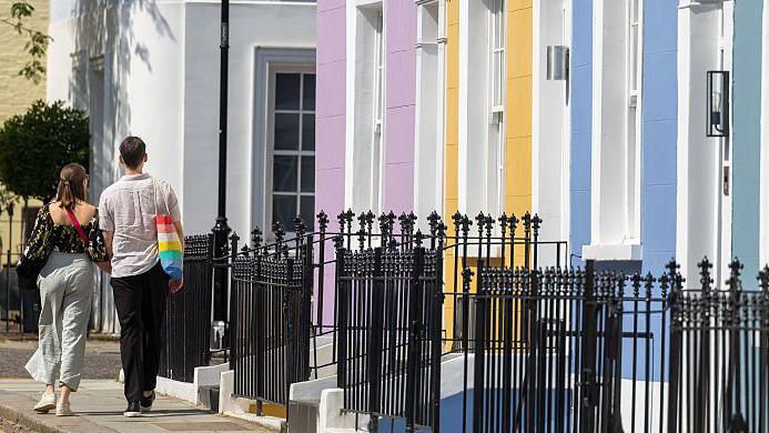 A couple photographed from behind walking down a residential street past houses painted pink, yellow and blue