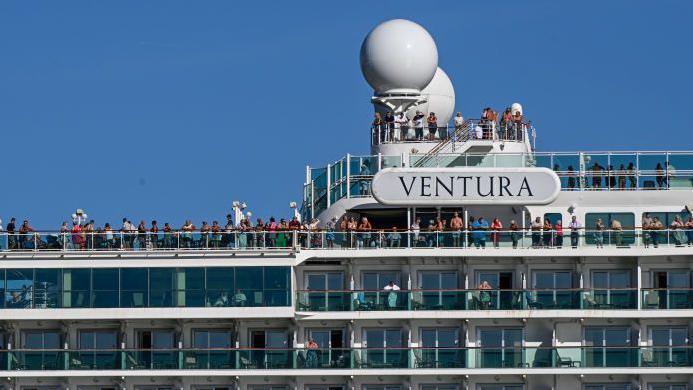 Close-up of passengers standing on the top deck of a cruise ship. A large name plate reading 'Ventura' is on the side of the ship. Cabin balconies can also be seen.