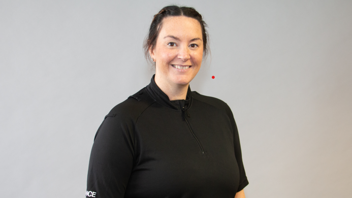 A woman with tied-back dark hair and a black police shirt smiles as she stands in front of a light grey wall.