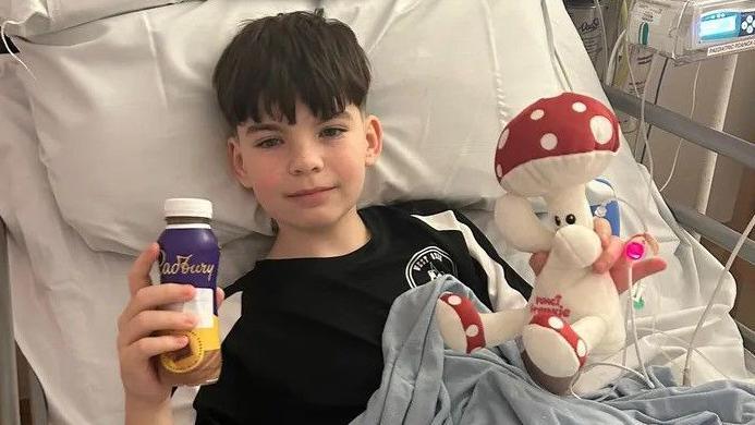 Josh in a hospital bed. He's holding up a Cadbury's milkshake bottle and a cuddly toy in the shape of a mushroom creature. He has black hair, thin on the sides, long at the front.