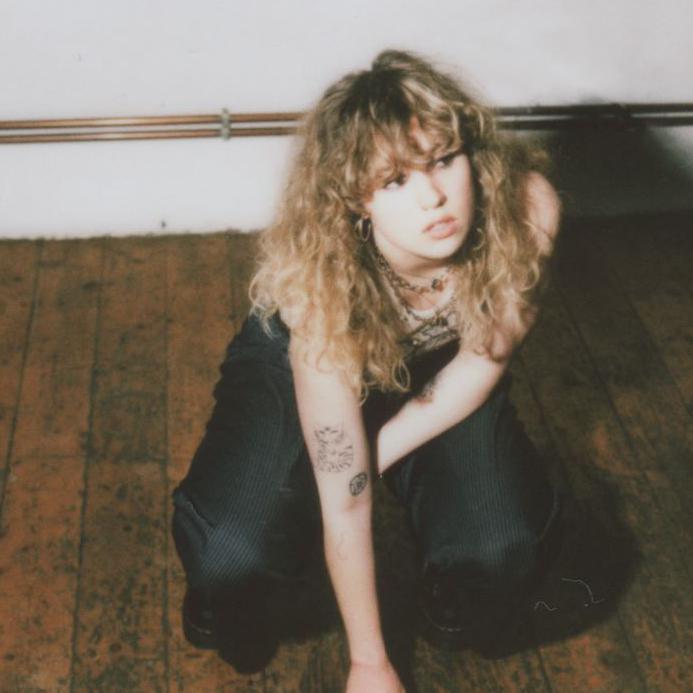 A young woman with blonde curly hair and a full fringe crouching on a wooden floor and looking to her left. She has a few small tattoos on her arm and is wearing lots of necklaces and black trousers.