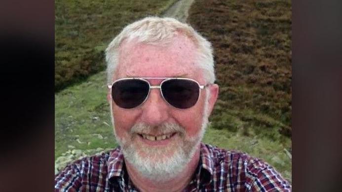Man with sunglasses and white beard and hair. He's wearing a shirt and you can see path on Great Hangman behind.
