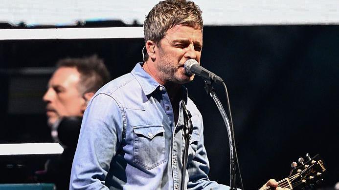 Noel Gallagher wears a denim shirt on stage while singing into a microphone. He scrunches up his face into the microphone. 