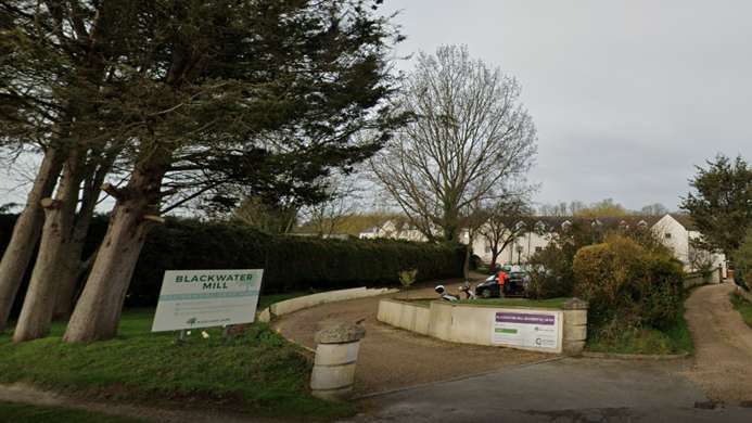 Exterior view of Blackwater Mill Residential Home - a white two-storey building at the end of a curved drive, with trees and a hedge to the left