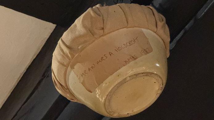 A cream ceramic bowl with brown fabric covering the top and a brown label that reads: "Mr an Mrs A Holdcroft, Xmas 1987"