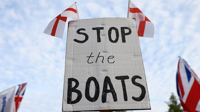 A white sign, with the words "Stop the boats" in black writing, and two small England flags attached to the top. There are union jacks being flown either side of the sign, which has the sky behind it.