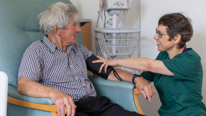 The picture shows a man and a nurse in a hospital setting.  The man is sitting in a chair having his blood pressure taken.