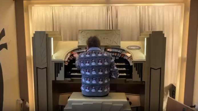 Mr Martin with his back to the camera  playing the organ, which is tucked into an alcove with a cinema-type curtain behind