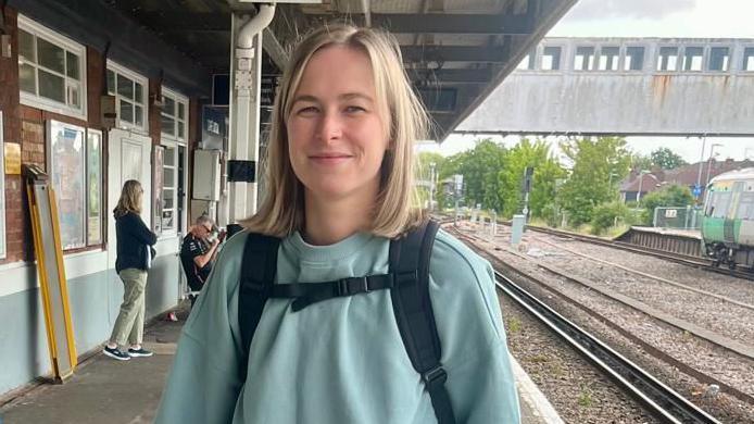 A woman with shoulder length blonde hair wearing a green jumper and a backpack standing at a train station with the railway track and a platform behind her. It is a head and shoulders shot of her.