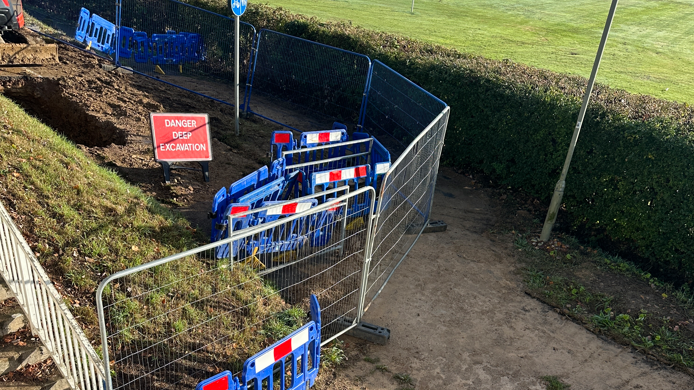 Metal and blue plastic barriers around a mound of earth with a large hole and playing fields to the left.