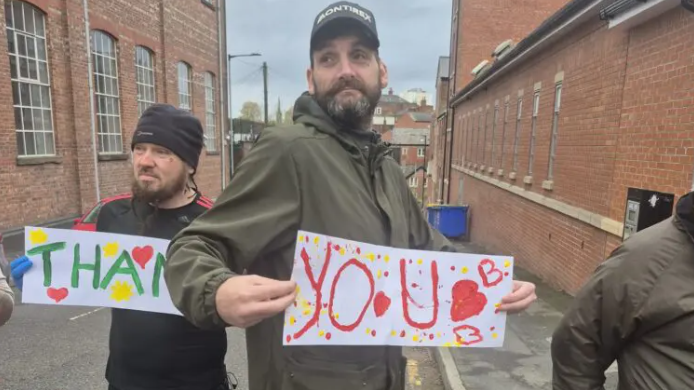 Several individuals are standing on a narrow street between two rows of red-brick buildings. The ground is wet, suggesting recent rain. Two people in the foreground are holding colorful handmade signs with large letters and heart decorations. One sign reads “THANK” and the other reads “YOU” with additional hearts and small drawings. The group is dressed in casual outdoor clothing, including jackets, caps and hats.