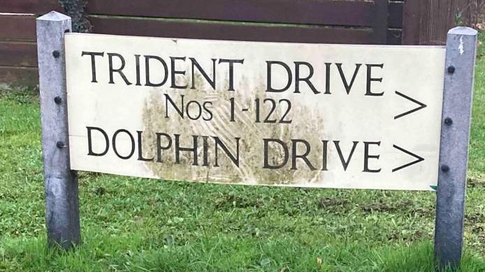 A rectangular white held up by two grey posts sitting in green grass. The words TRIDENT DRIVE Nos 1- 122 and DOLPHIN DRIVE are written in black on the sign.