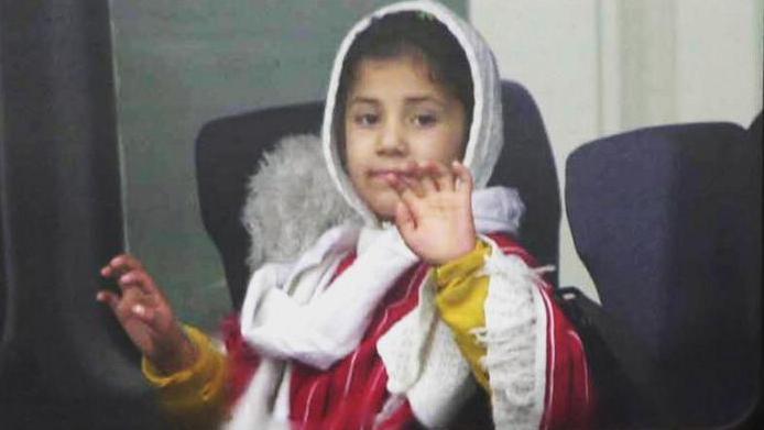 A child, wearing a white hood and red robe, waves from a bus after leaving the aircraft