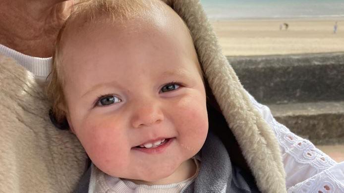 A baby smiles at the camera, while sitting on a woman's knee.