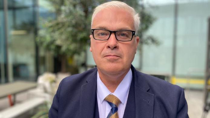 Michael Hudson is looking directly at the camera, unsmiling. He is wearing a blue three-piece suit, with yellow and blue tie. He has black glasses and white hair. He is stood in a glass atrium.